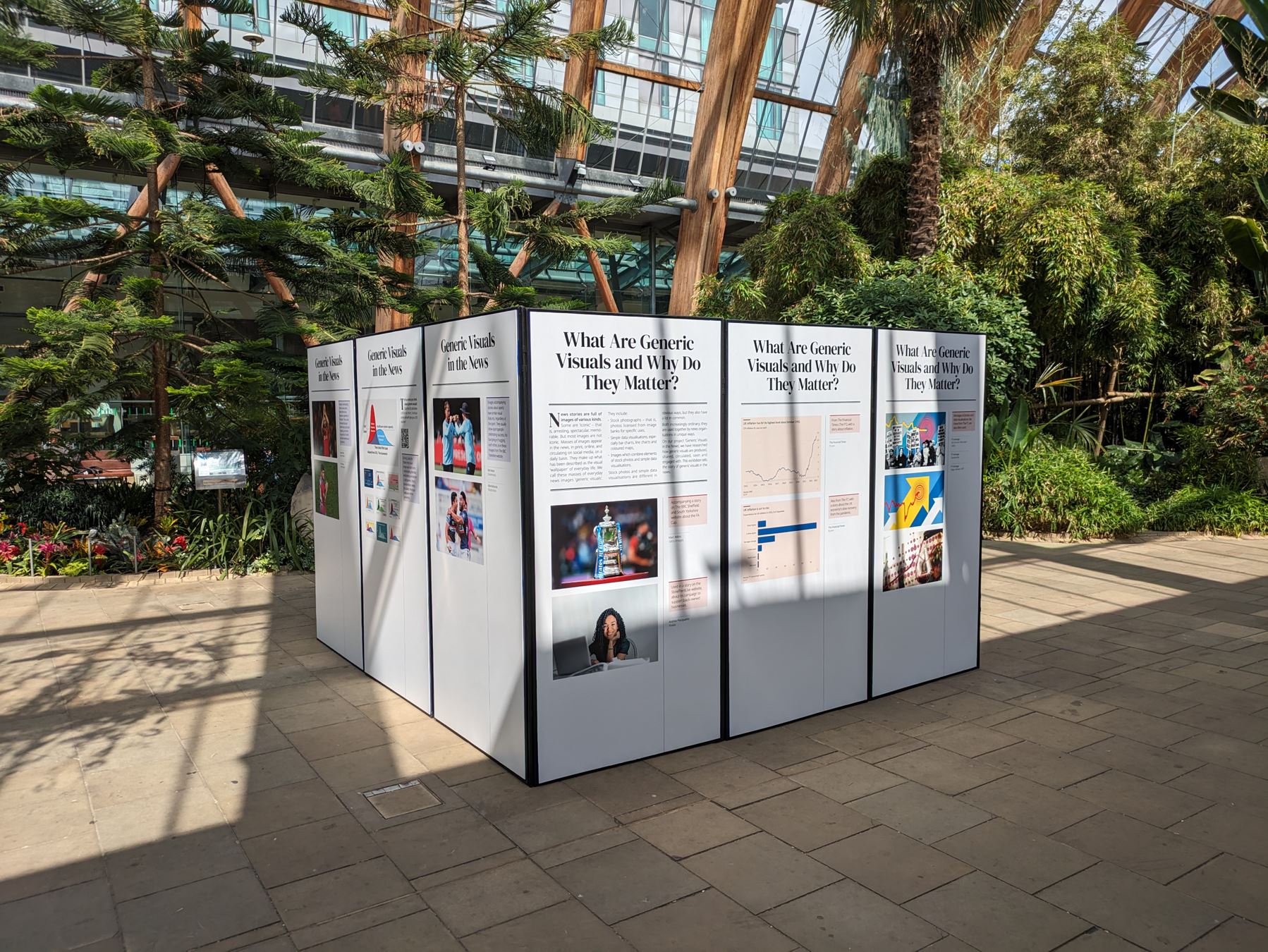 Image is a photograph of the Generic Visuals in the News exhibition installed in its location at the Winter Garden in Sheffield city centre. The exhibition is displayed on a series of white panels arranged in a square-like configuration. The backdrop is of vegatation; trees and the roof of the hothouse. There is a shadow cast onto the exhibition panels.